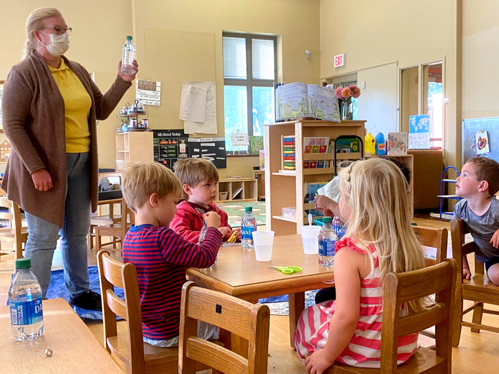 Dr. Betty Lise Anderson stands in front of preschoolers sitting at child-sized tables. She is holding up a bottle of water as the kids look on.
