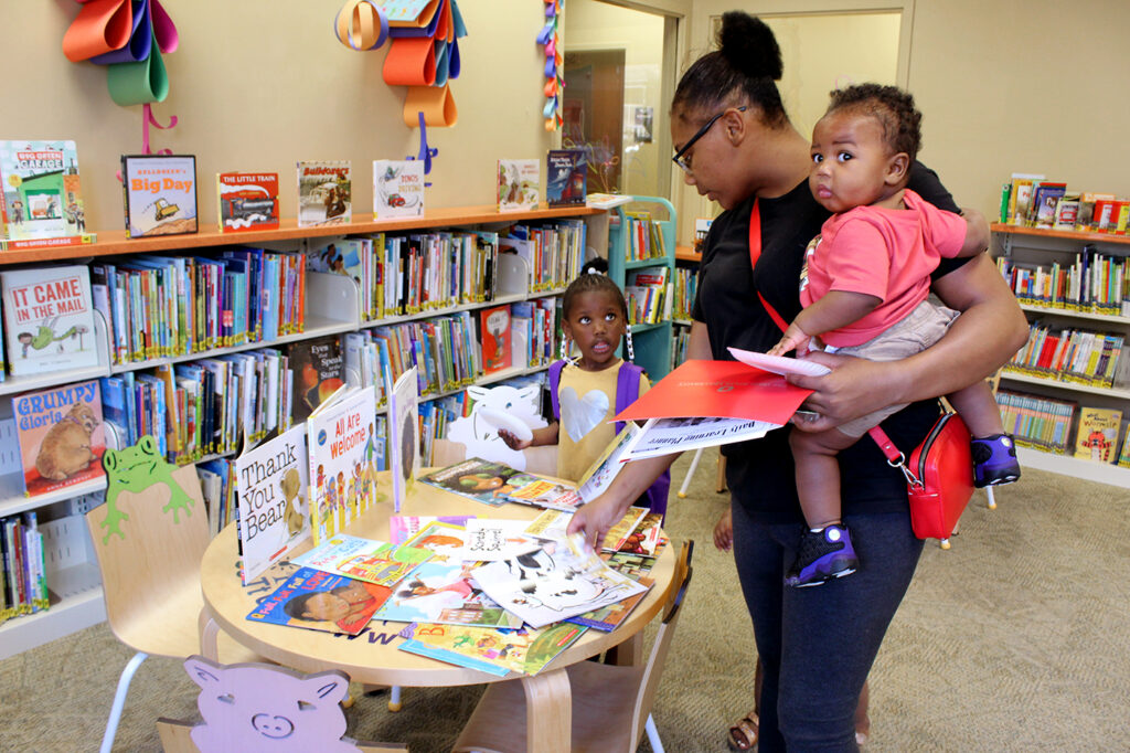 Mom looks at books on a table in the library while holding a baby while another child stands looking at Mom.