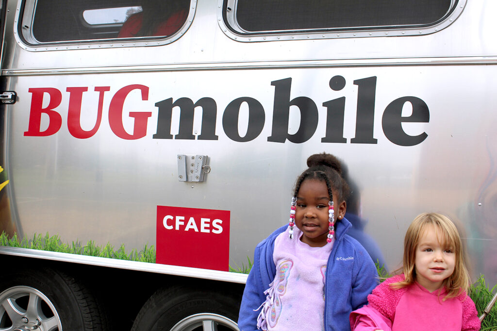 Two children pose for a picture outside of the BUGmobile, a converted silver RV with the logo on the side.