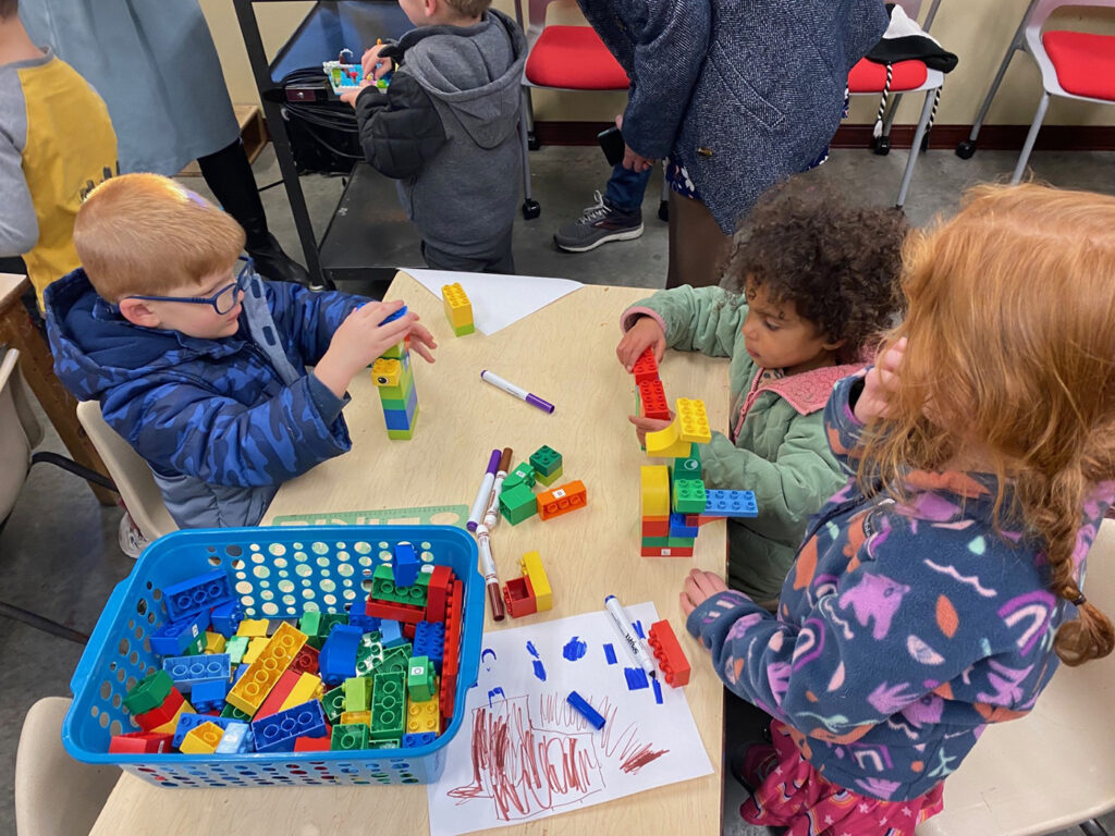 Three small children around a rectangular table construct towers out of Lego blocks.