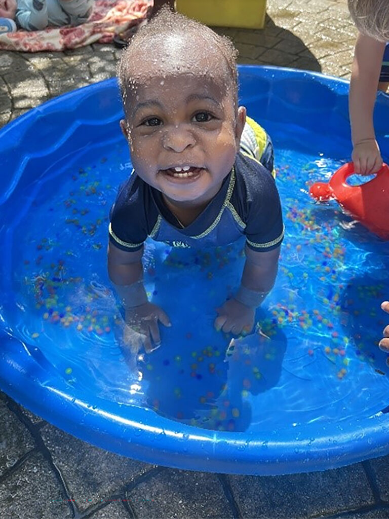 A child crawls in a small wading pool containing two inches of water. The child is wearing a blue shirt and is looking at the camera and smiling.