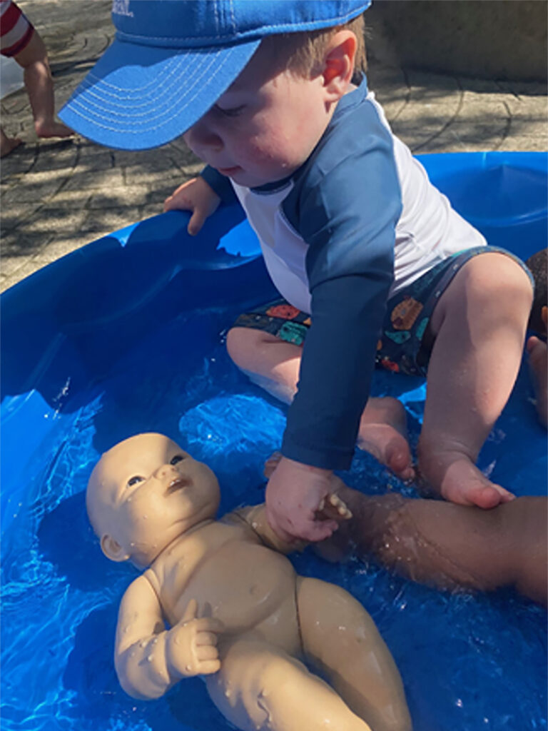 A young child sits in about two inches of water in a small wading pool. The child holds the hand of a baby doll that is lying face up in the wading pool.