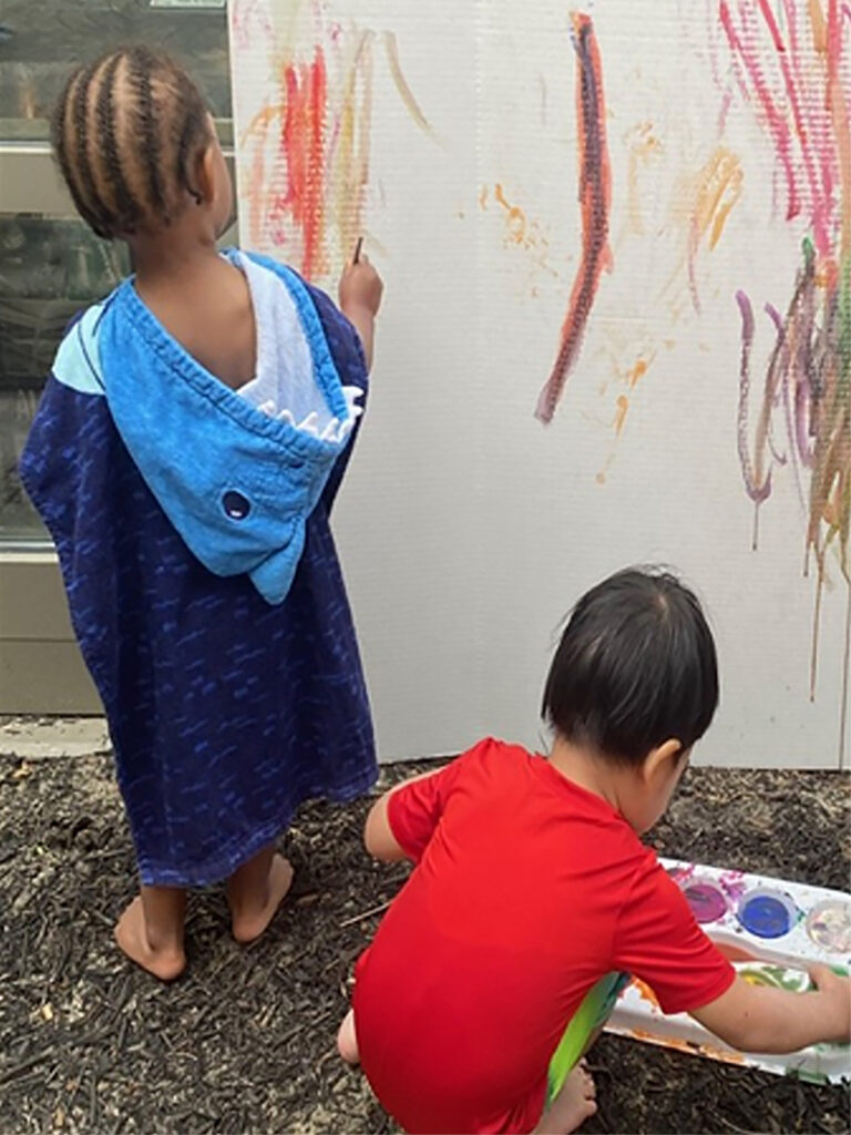 Two children play with watercolor paints outside. One child holds a brush and paints a display board propped against a wall, while the other child crouches over a plastic tray on the ground with round indentations containing different colors of paint.