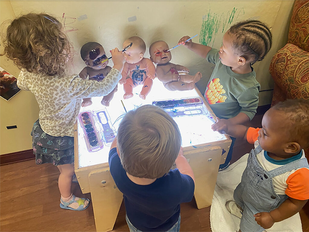 A group of small children dab watercolor paint of various colors onto baby dolls. The children stand around a small table with a lightboard surface. Two trays of watercolor paint are on the table, along with three dolls propped up in seated position against a wall at the back of the table.