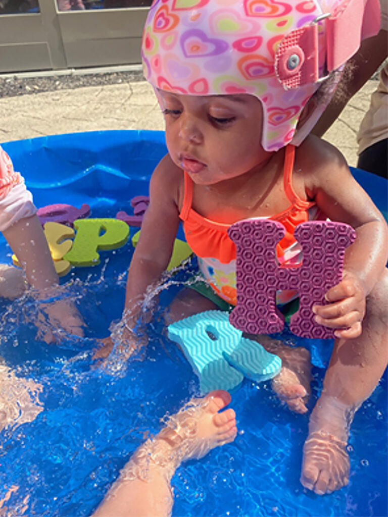 A small child sits in about two inches of water in small wading pool holding a foam cutout of the letter "H." Floating in the pool are cutouts of other letters of the alphabet. The child is wearing a pink helmet-style cap with hearts printed on it. The hands and feet of other children in the wading pool are also visible.