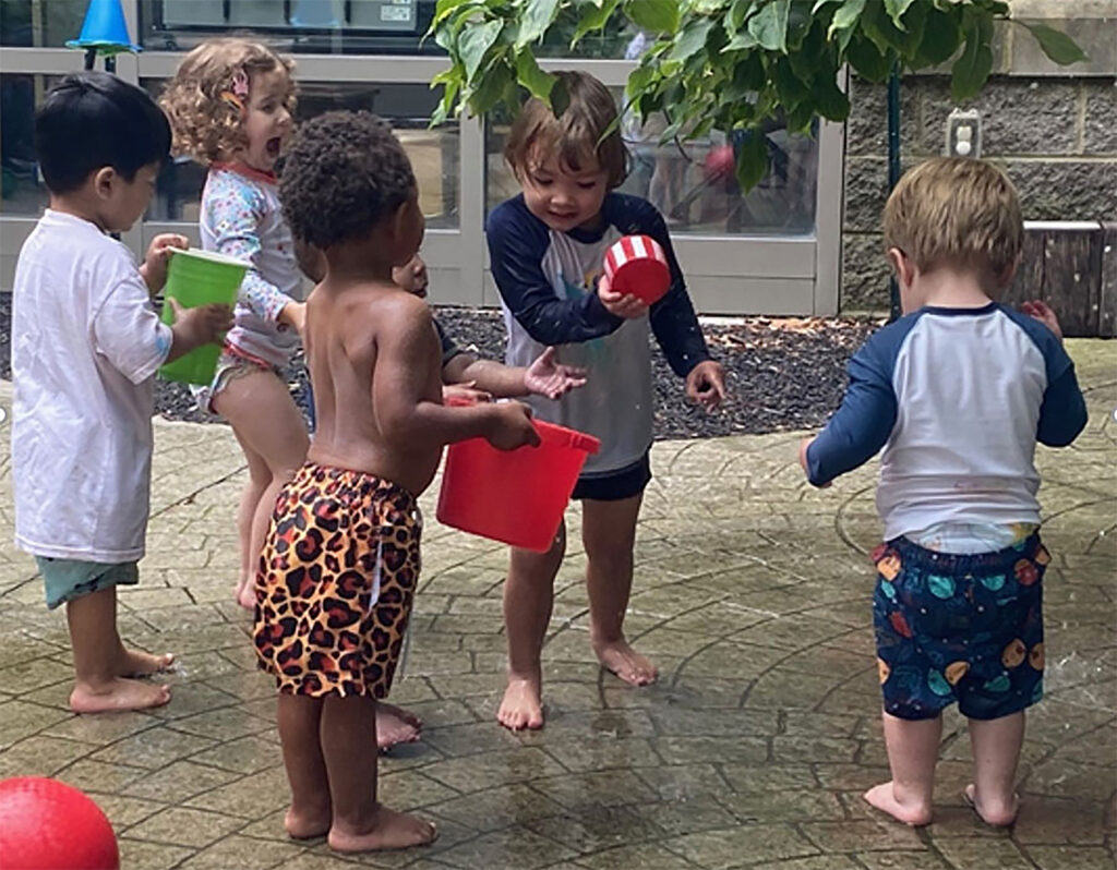 Six small children are standing barefoot on a stone-paved patio holding cups and small containers with water.
