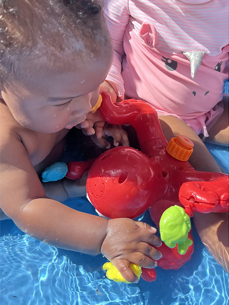 A young child plays with a red plastic octopus-style creature in a small wading pool. The child's hands are on two of the toy's arms. Another child sits nearby on the edge of the wading pool with a hand on one of the toy's arms.