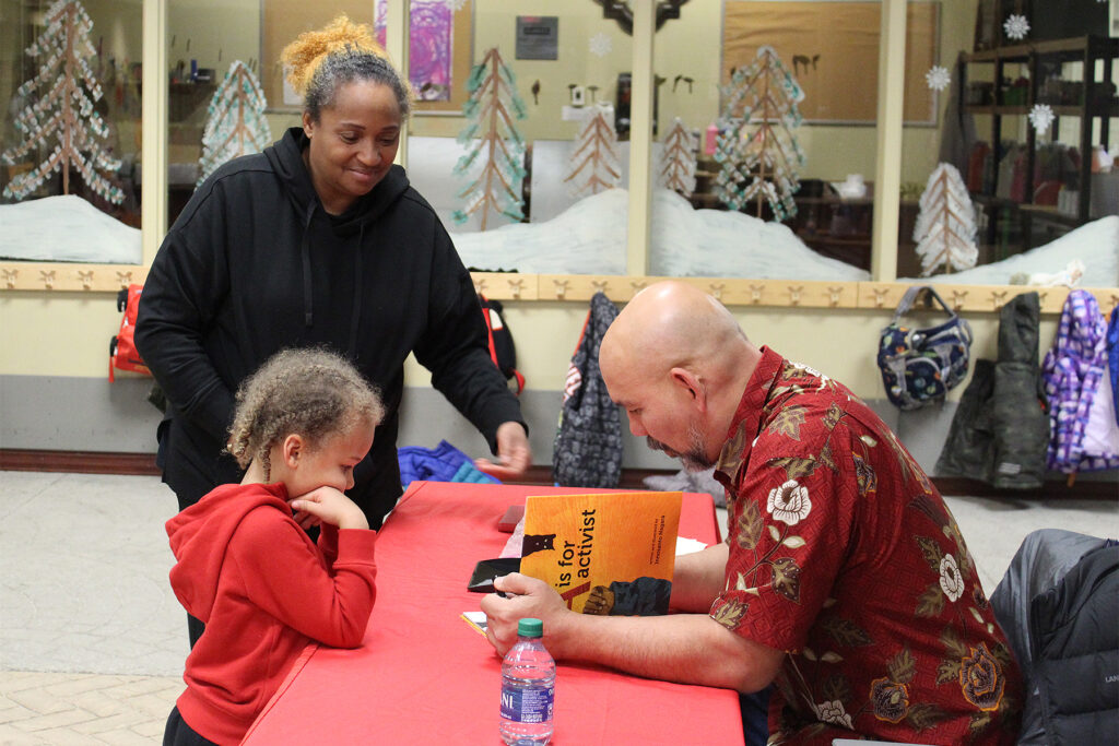Author Innosanto Nagara sits at a table on the right, signing a copy of his book "A Is for Activist," as a young child stands across from him watching. Standing to the child's left is an adult woman with her left arm extended to receive the book when Nagara finishes signing it.
