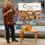 Innosanto Nagara stands, holding a microphone in his right hand and one of the books he has written, "M Is for Movement," in his left hand, as he speaks at the Schoenbaum Family Center. Behind him is a poster that reads "C is for our school community," and next to him is a small table with copies of other books and a small globe.
