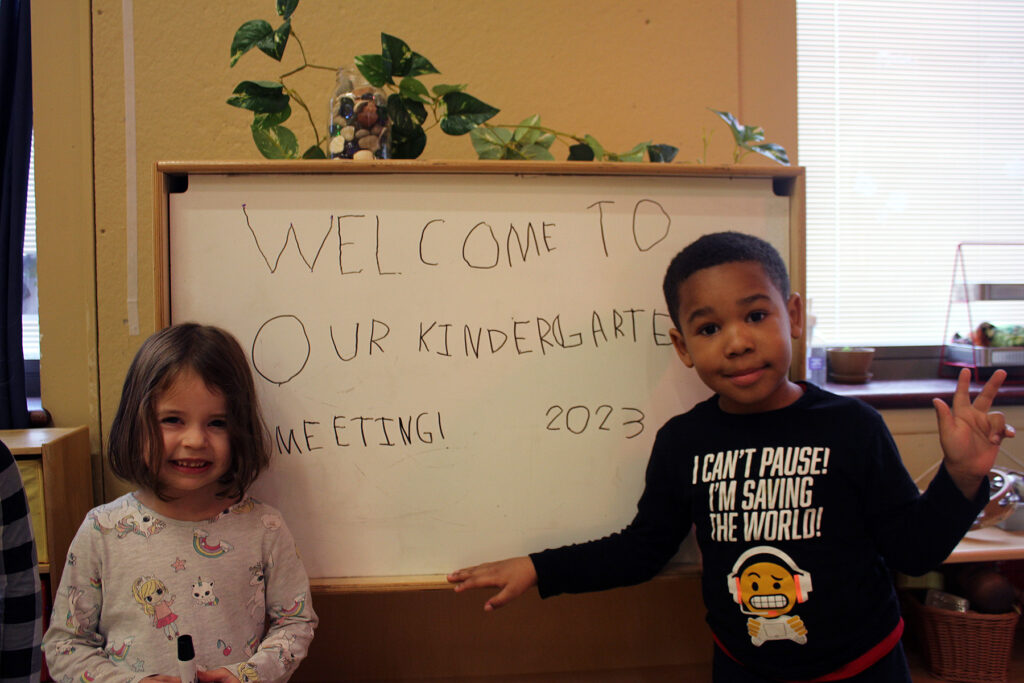 Two children stand next to a whiteboard on which is written in a child's block printing "WELCOME TO OUR KINDERGARTEN MEETING! 2023"