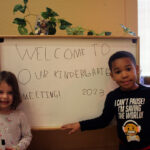 Two children stand next to a whiteboard on which is written in a child's block printing "WELCOME TO OUR KINDERGARTEN MEETING! 2023"