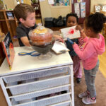 Three small children surround a storage cart on top of which is a glass terrarium containing a decaying pumpkin. Also on top of the storage cart is the book "Pumpkin Jack."