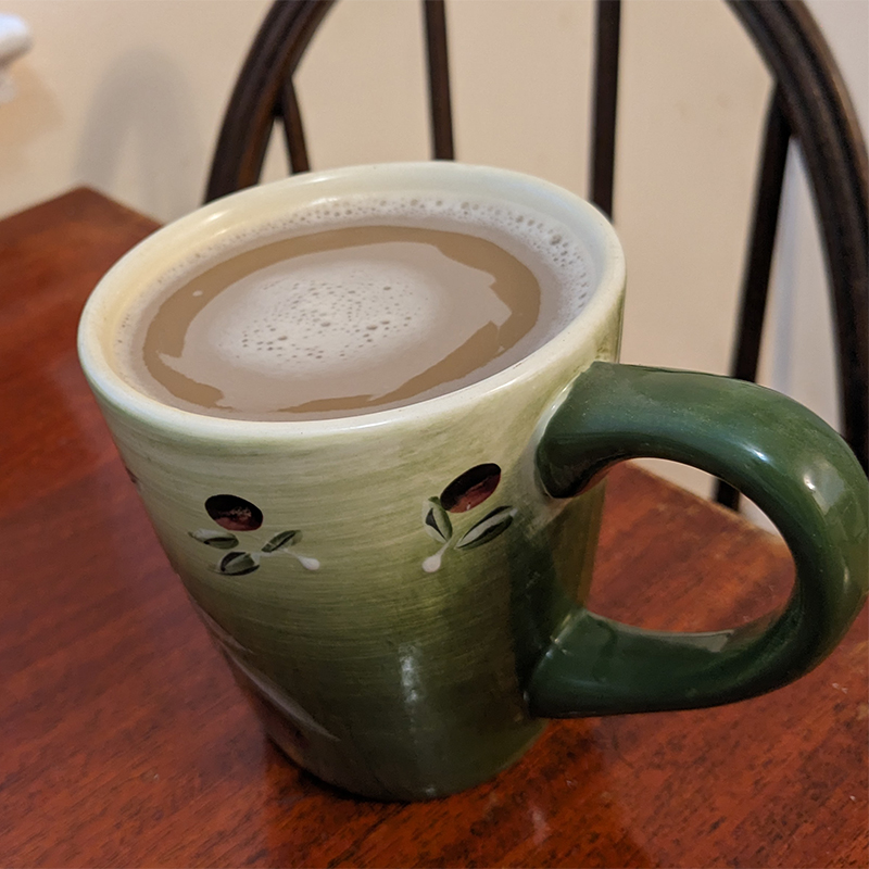 A large green mug filled with coffee rests on a wooden table. The back of a wooden Windsor chair can been seen in the background.
