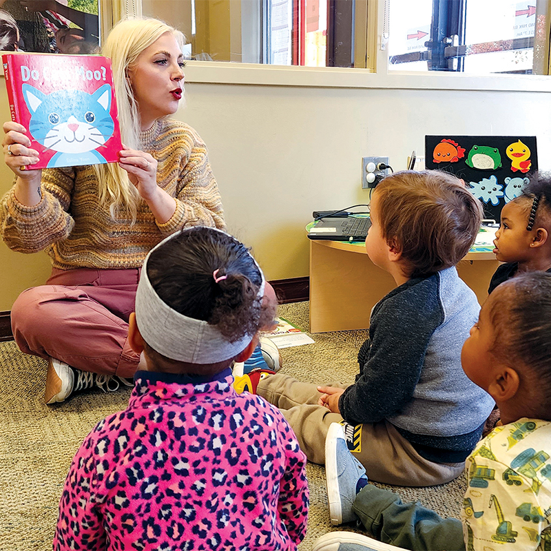 A woman sitting on the floor holds up a children's book titled "Do Cats Moo?" as young children who are also sitting on the floor watch her.