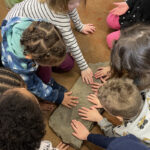 Young children sitting on a carpeted floor place their hands on a flat rock that contains footprints of dinosaurs.