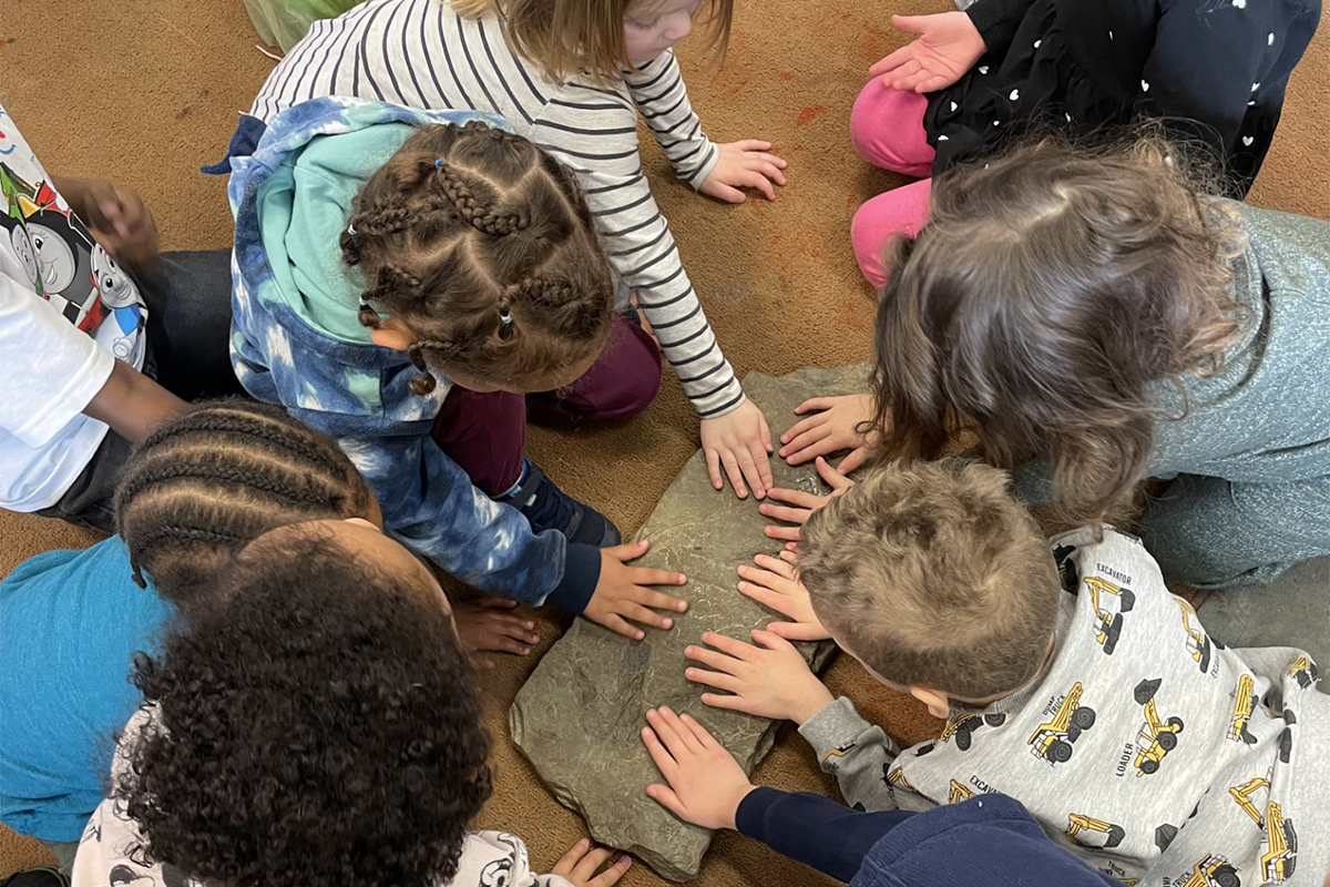 Young children sitting on a carpeted floor place their hands on a flat rock that contains footprints of dinosaurs.