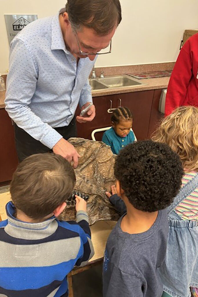 A man standing in back of a table points to fossils of dinosaurs in a rock on the table as young children in front of the table place their hands on the rock.