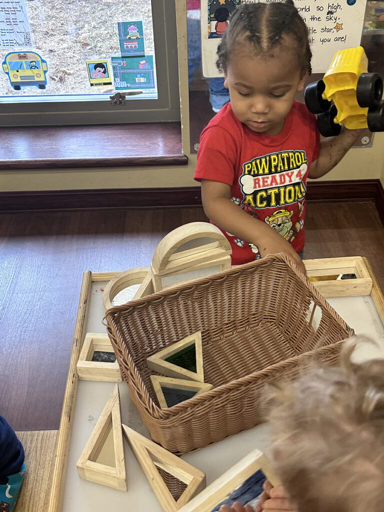 A small child holds a small, plastic toy truck in the left hand while looking over a small table and wicker basket containing wooden blocks of different sizes and shapes.