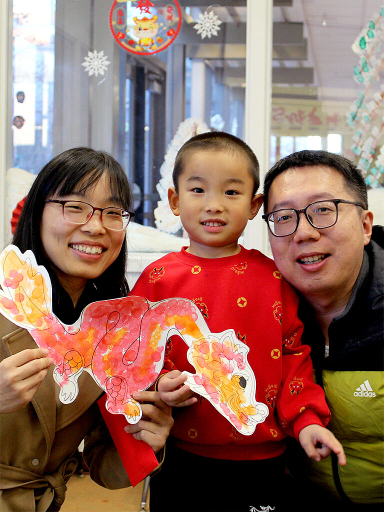 A small child stands between two crouching adults, so that the heads of all three are on the same level. On the left, a woman and the child hold a piece of paper on which a drawing of a dragon has been colored in orange and pink. A man wearing glasses is on the right.