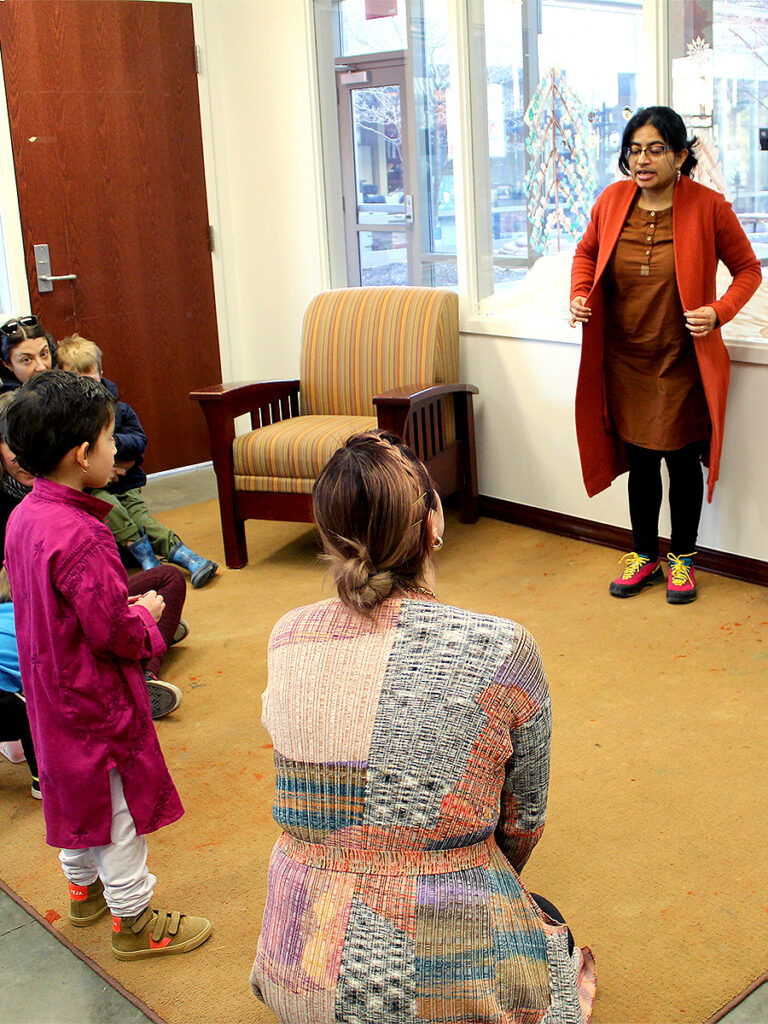 A woman stands on the edge of an area rug on one side of a room, facing a small child standing on the opposite edge of the rug. Other families are seated on the rug listening to the woman.