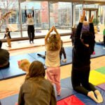 A yoga instructor, standing on a mat, lifts both hands into the air, as adults and children try to repeat the pose either standing or perched on their knees with their backs erect.
