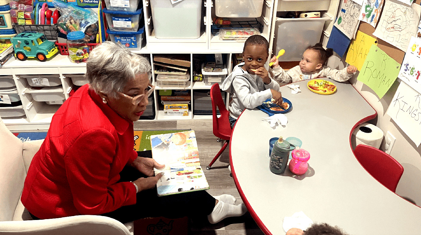 U.S. Representative Joyce Beatty, left, sits in a chair and reads a children's book to several children eating meals on colorful plates at a small table.