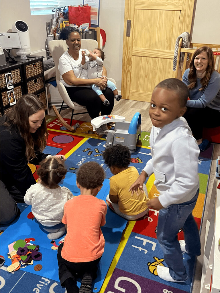 A small child turns and looks at the camera as other small children sit on the floor looking toward a woman seated in a rocking chair holding an infant.