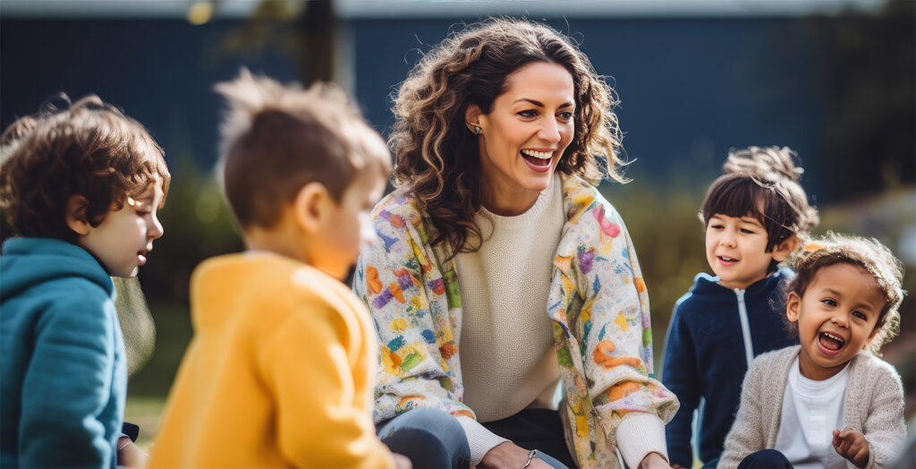 A teacher smiles surrounded by smiling small children outdoors during a play activity. The preschool-age children are wearing jackets, as is the teacher.