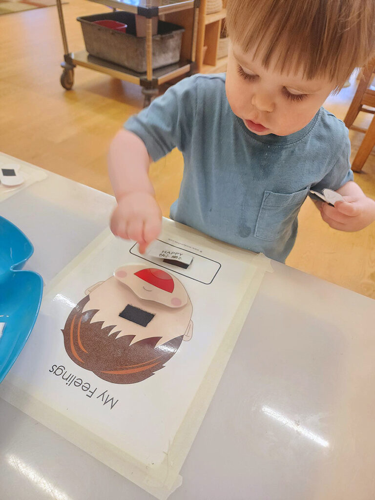 A small boy places a card depicting a smiling, wide-open mouth on a blank, cartoon face taped to a table. Above the face are the words "My Feelings."
