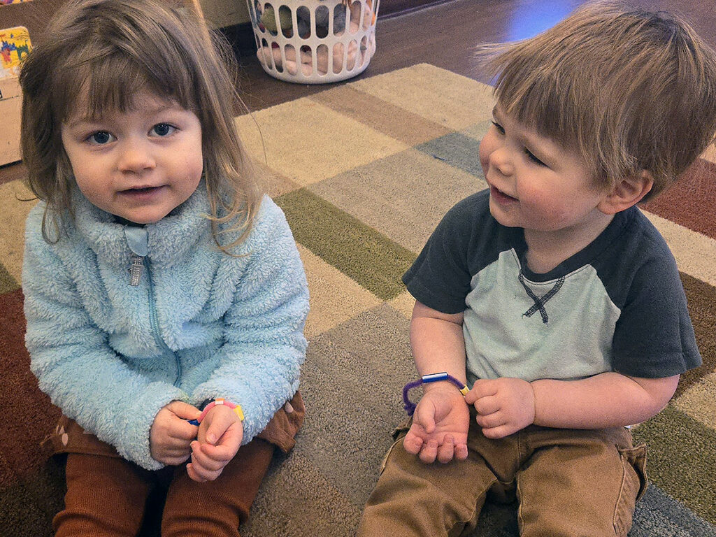 A small boy, right, with a wrist band on his right hand, looks at the face of a small girl, left, who has a wrist band on her left hand and is looking at the camera.