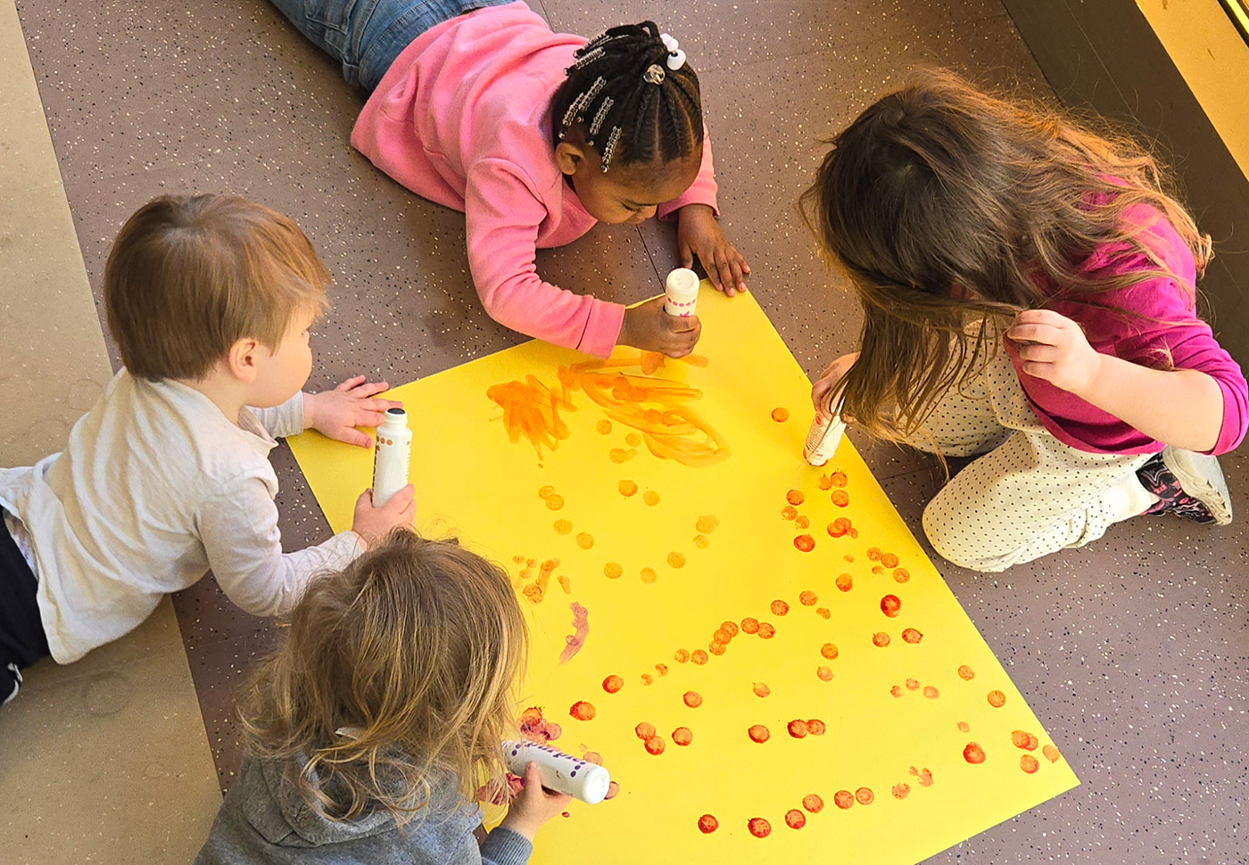 Four small children surround a large sheet of yellow construction paper, stamping red circles on the paper using large ink stamps.