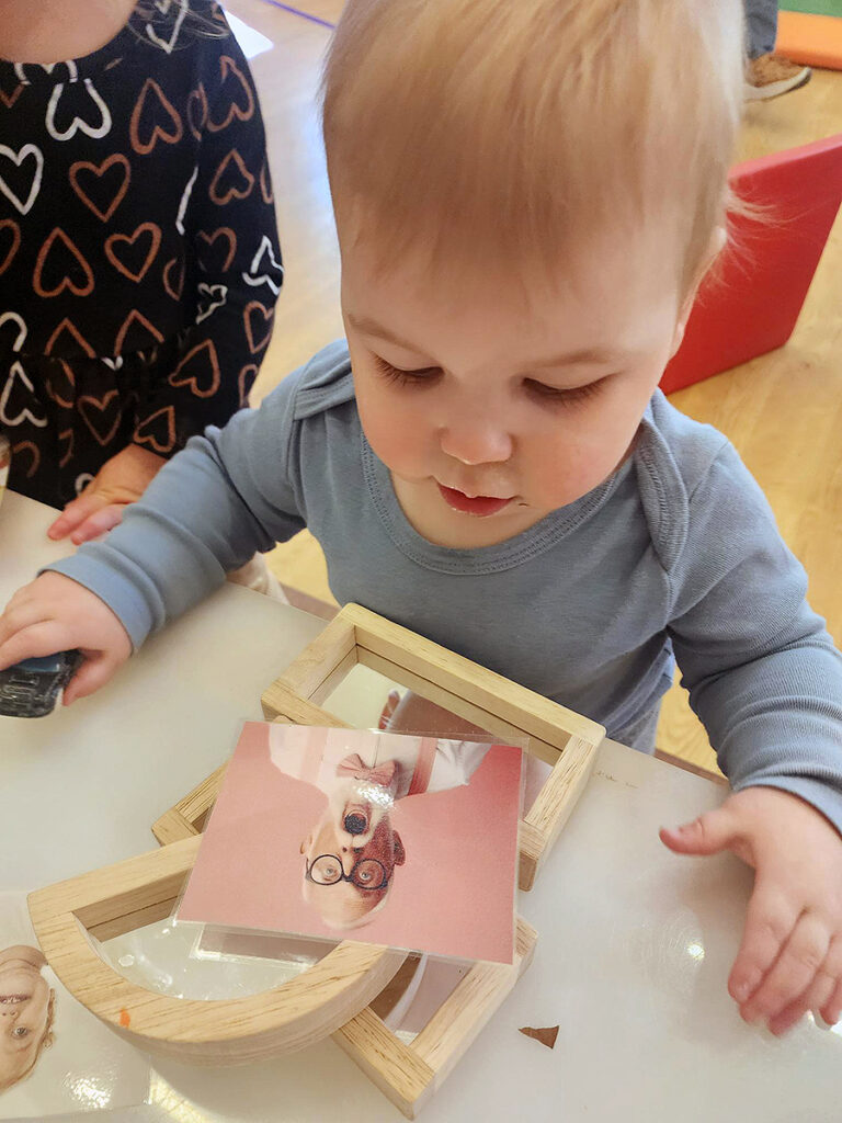 A toddler looks at the photograph of an older man with glasses, a white mustache and goatee. The man's mouth is open in a large circle, and the man's eyelids are open wide.