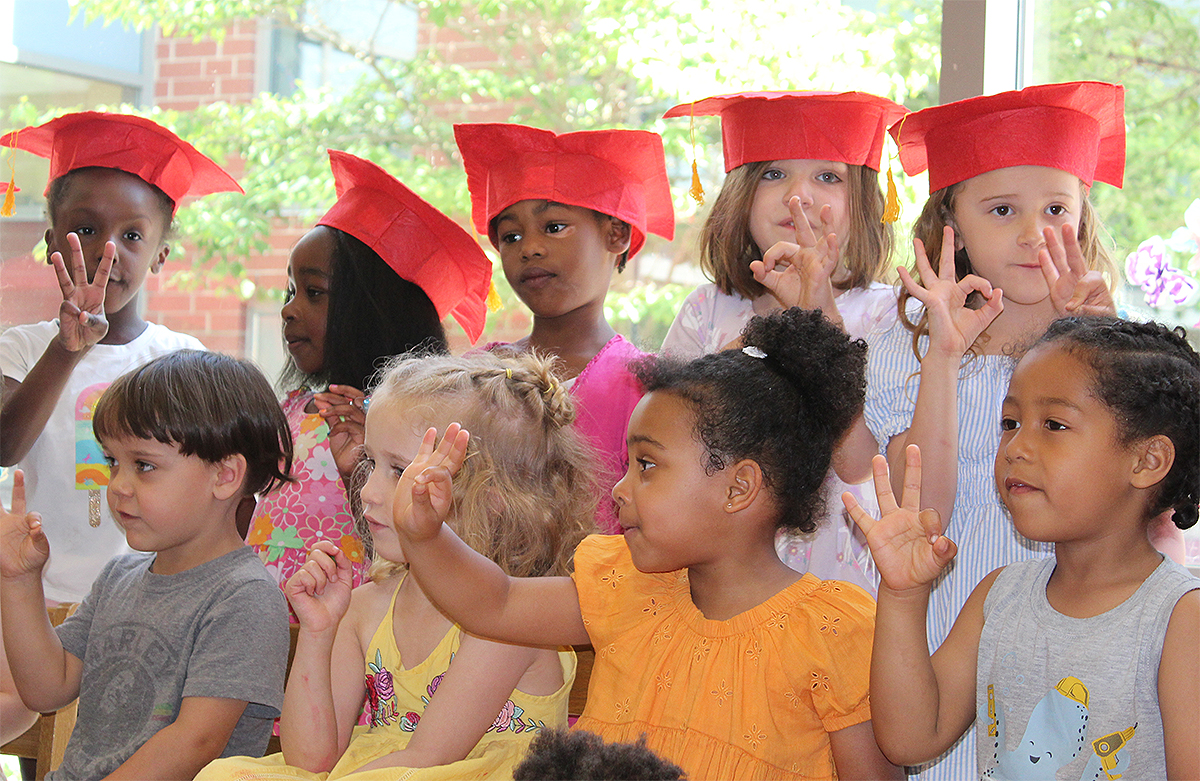 A group of young children, some wearing scarlet-colored mortar-board hats, hold up three fingers on one hand.