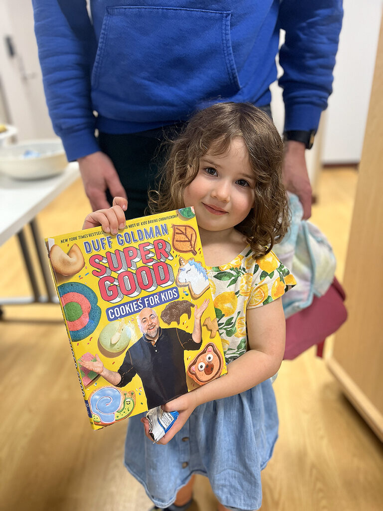 A young girl holds up a copy of the book "Super Good Cookies for Kids" by Duff Goldman.
