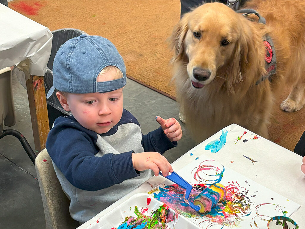 A golden retriever watches a young boy seated at a small table as the boy paints on a piece of paper using a roller with different grooved shapes. The boy is wearing a baseball cap turned back side front. The piece of paper is covered with a variety of bright colors from the paints.
