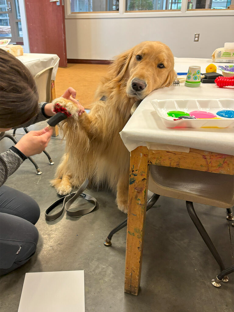 A golden retriever holds up its right front paw, the underside of which is being painted red by a woman. A sheet of white paper is on the floor, onto which the paw will be pressed to leave a red impression. The dog's head is leaning on a table on which is a styrofoam container with chambers holding various colors of paint.