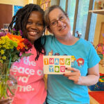 A. Sophie Rogers teachers Luanda Cunningham, left, and Ashley Keener stand together smiling, each with an arm behind the back of the other. Luanda holds a large glass vase in her right hand, and the vase is filled with a variety of colorful flowers. Ashley holds to face the camera a card on which is written "THANK YOU."