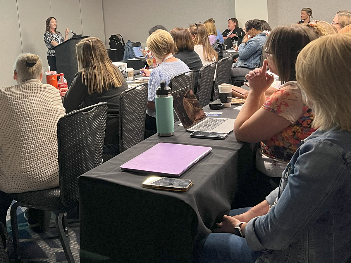 A woman stands at a podium in a crowded conference room and speaks into a microphone on the podium. Members of the audience sit in rows of long tables taking notes with pen and paper or laptop computers as the woman speaks.