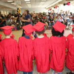 A group of preschool children wearing red graduation gowns and red mortar-board hats are seen from the back facing an audience of parents and family members cheering them on. Many of the family members in the audience are holding up cell phones to take pictures, and colorful balloons adorn the ceiling of the large room.