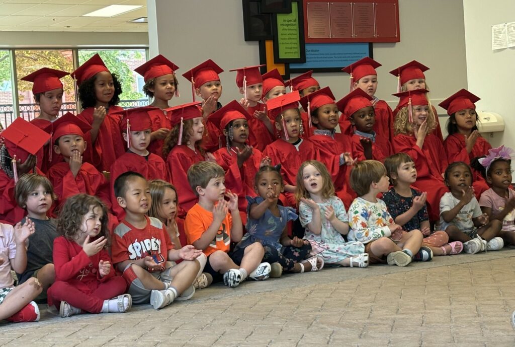 Preschool students wearing red graduation gowns and red mortar-board hats form two rows as they face the audience. In front, sitting on the floor, is a row of other students who are not wearing the cap-and-gown.