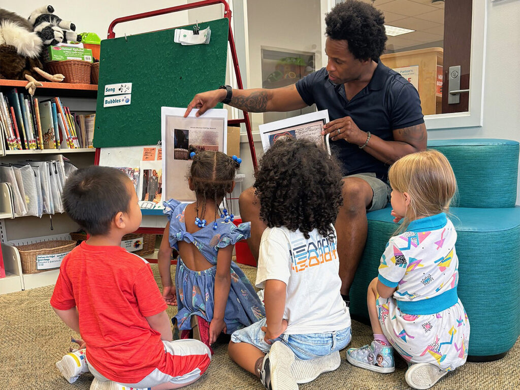 A man sits on an upholstered seating area in a preschool library holding printouts of photos of items of family history to a group of small children.