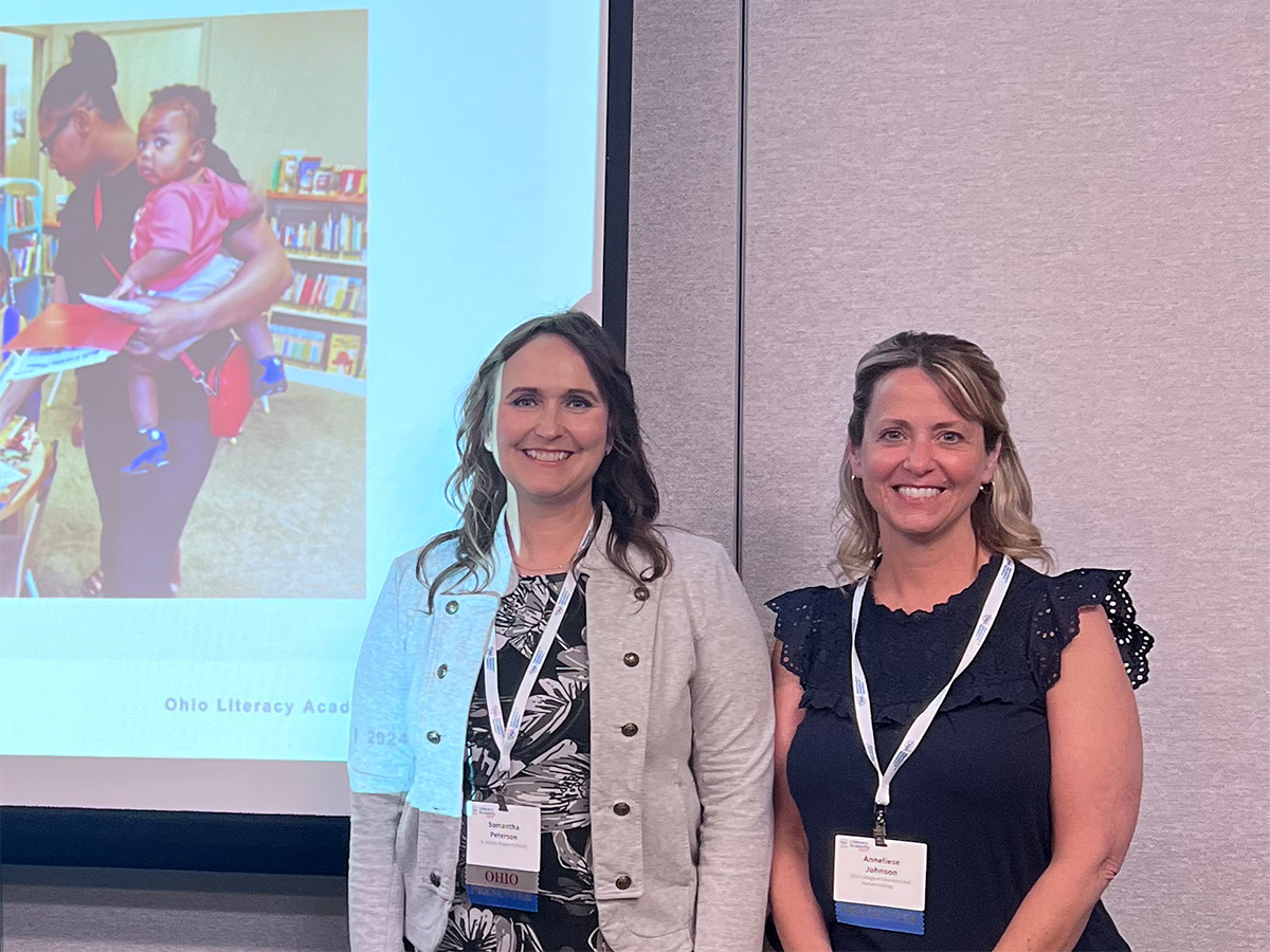 Two women face the camera, standing in front of a wall and a screen. The women are wearing lanyards identifying them as speakers at a conference. On the screen is a projected image showing a woman holding a small child aloft tucked in her left arm while the two are inside a preschool classroom.