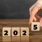 Four small wooden blocks are lined up in a row, with the fourth block being put in place by a hand holding it between the thumb and forefinger. On the front face of each block is a number, so that when placed together the blocks read 2025.