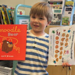 A young child holds up the book "Noodle Bear" by Mark Gravas in his right hand, and a sheet of paper for the ASR Summer Literacy Challenge in his left hand. The child, who has streaks of paint on the face and is wearing a blue-and-white horizontally striped shirt, is standing in a library in front of carts of books and brochures.