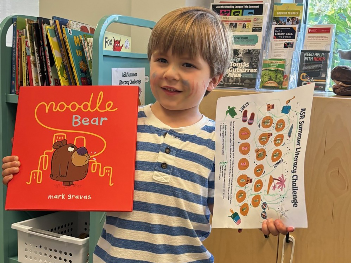 A young child holds up the book "Noodle Bear" by Mark Gravas in his right hand, and a sheet of paper for the ASR Summer Literacy Challenge in his left hand. The child, who has streaks of paint on the face and is wearing a blue-and-white horizontally striped shirt, is standing in a library in front of carts of books and brochures.