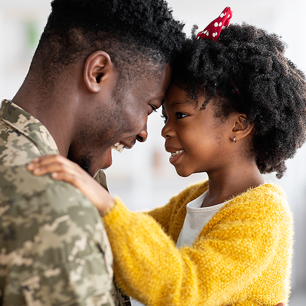 A man wearing camouflage military fatigues is next to a young girl in a yellow sweater. The man and the girl smile at each other with their foreheads touching.