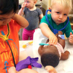 Two toddlers play doctor using baby dolls on a table in a classroom. There is a young boy in an orange shirt putting on a stethoscope, while the second boy in a teal shirt pushes the stethescope onto the chest of a baby doll on the table.