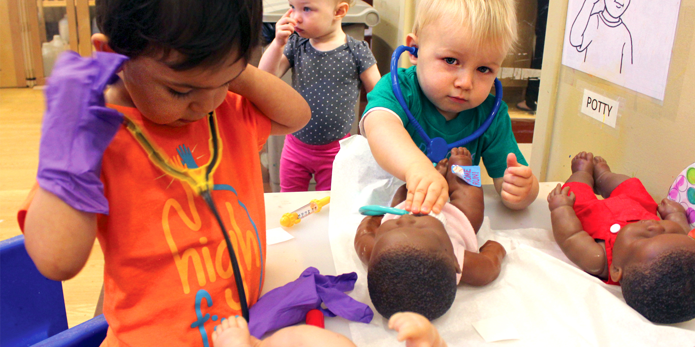 Two toddlers play doctor using baby dolls on a table in a classroom. There is a young boy in an orange shirt putting on a stethoscope, while the second boy in a teal shirt pushes the stethescope onto the chest of a baby doll on the table.