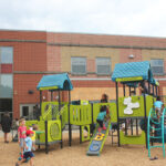 Several small children play on a playground. Dominating the playground is a giant green structure with blue roofs covering several ladders, climbing areas, slides and play areas.