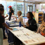 Three people sit behind a long table with various flyers and brochures in front of them.